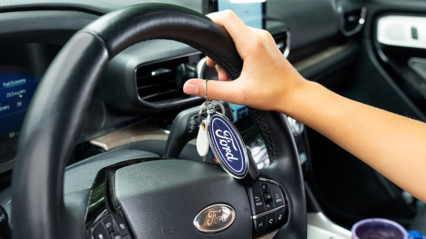 Hand on a steering wheel holding an oval keychain customized with a Ford logo
